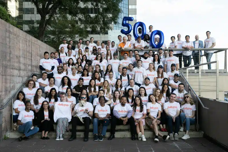 A large group of people wearing matching "daily pay" T-shirts poses for a photo on outdoor steps. Balloons forming the number "500" are held up in the background.
