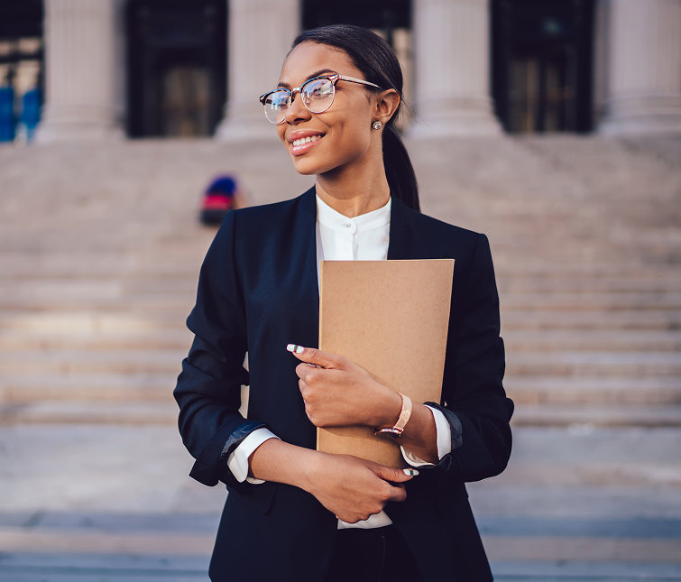 A woman in business attire stands outside a building with columns, smiling and holding a brown folder.