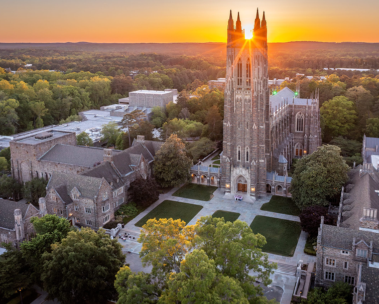 Aerial view of Duke University Chapel at sunset, with sunlight shining through the tower and surrounding campus buildings and trees visible.
