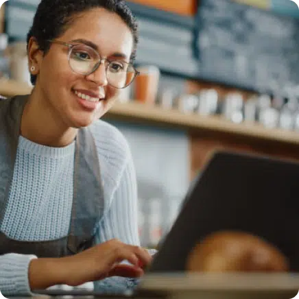Person with glasses and an apron smiling while using a laptop in a cozy setting.