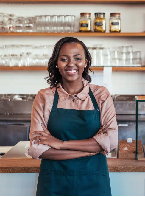 A person in a green apron and pink shirt smiles behind a counter with glasses and jars on shelves in the background.