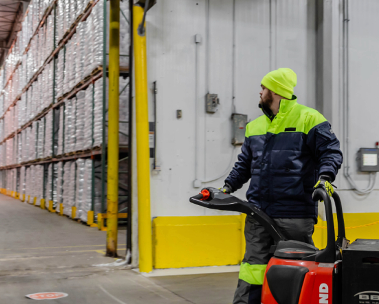 A worker in a high-visibility jacket and beanie operates a pallet jack in a large warehouse with stacked goods.