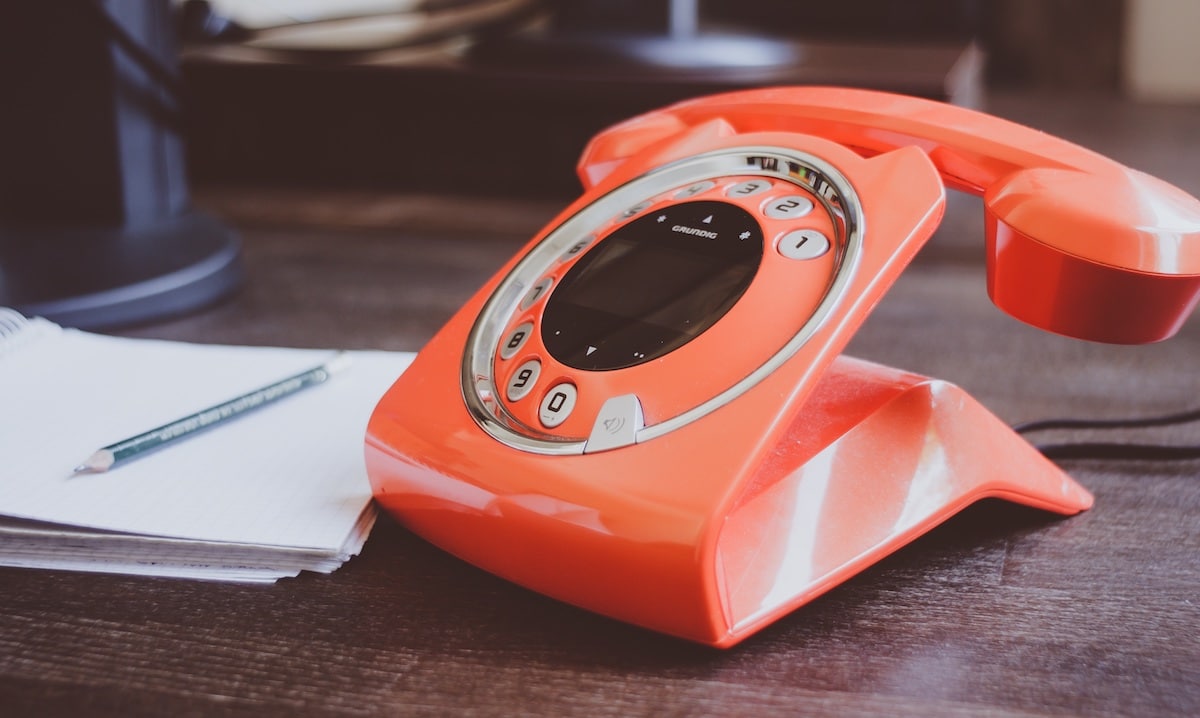 A vivid orange retro-style telephone with a digital display rests on a wooden desk beside an open notebook and a pencil.