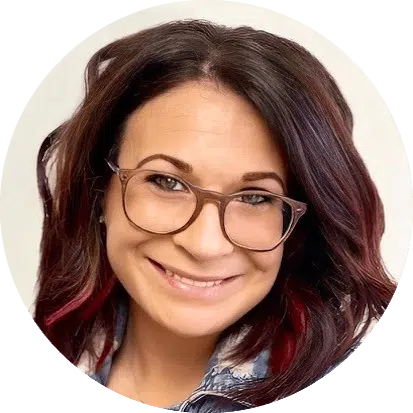 A person with shoulder-length dark hair, red highlights, wearing glasses, and a denim jacket, smiling at the camera.