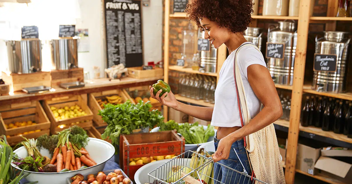 A young woman with curly hair wearing a white t-shirt and beige tote bag smiles as she holds a green bell pepper in a grocery store. She stands near bins filled with various vegetables like carrots, lettuce, and turnips. Shelves with jars and a blackboard with handwritten notes about On-Demand Pay benefits are in the background.
