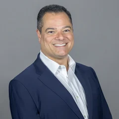 A man in a navy blue suit and white shirt smiles at the camera against a plain gray background.
