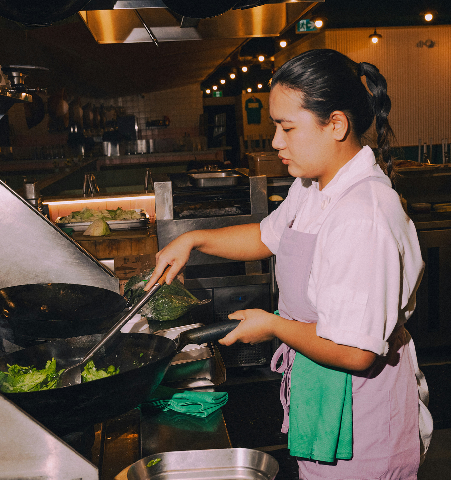 A chef wearing a white uniform and apron stir-fries green vegetables in a wok in a restaurant kitchen.
