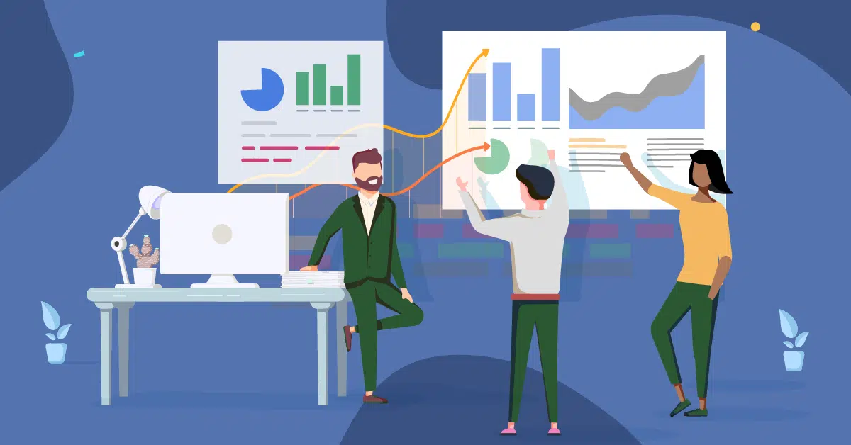 Three people in an office discuss charts and graphs displayed on a wall while one person sits on a desk and two others point at the charts.