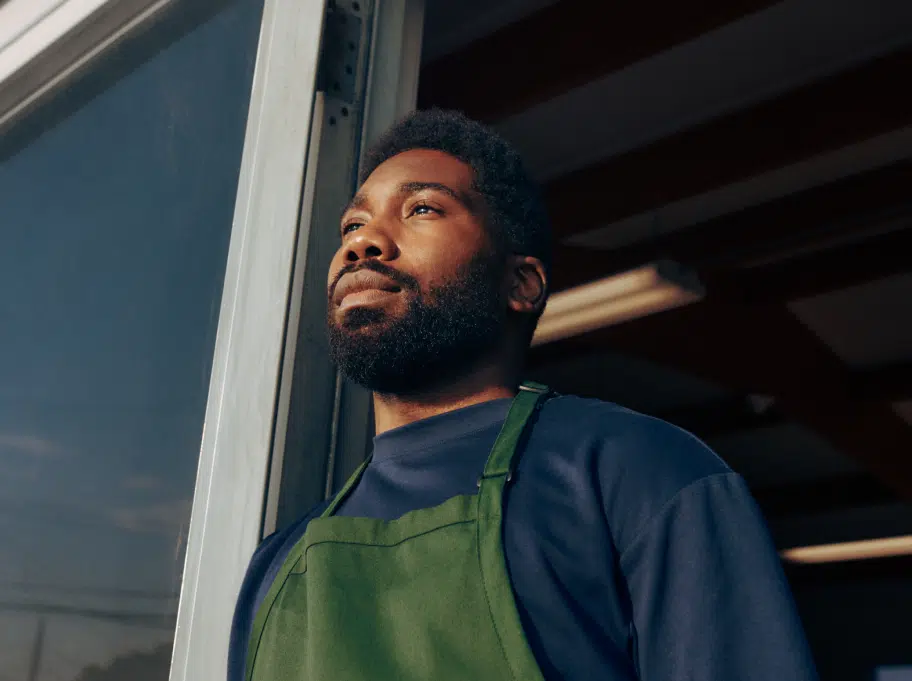 A person with a beard and short hair wearing a green apron and dark blue shirt stands near a door and gazes upwards.