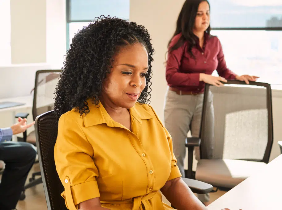A woman in a yellow shirt is working at a computer desk, while another woman in a maroon shirt stands in the background adjusting a chair in a brightly lit office.